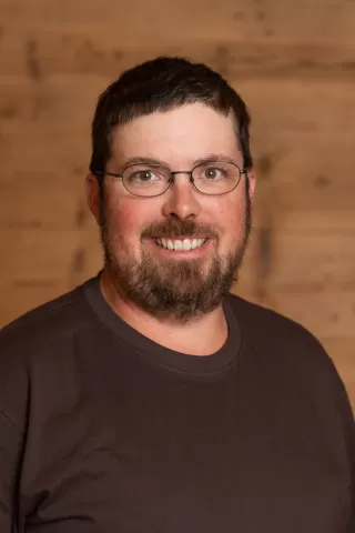 Mitarbeiterportrait / Foto mit braunem T-Shirt oder schwarzem Polo Shirt mit Zimmerei Hirschi Logo. Strahlend und lachend nach oben schauend oder direkt in die Kamera.